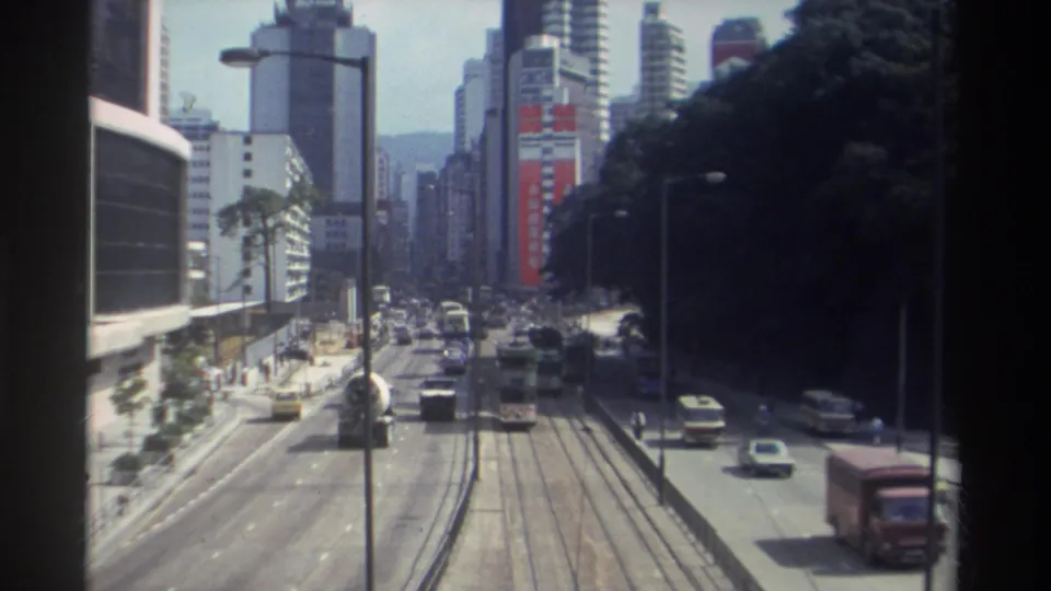 1982 Hong Kong street seen from above, with tram rails, moving traffic, buses, tall buildings, and a dark wooded hillside at right.
