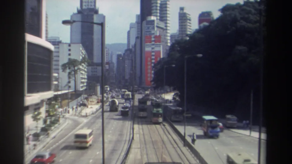 Hong Kong street traffic in 1982, with a bus approaching, cars moving in several lanes, central tram tracks, and high-rise buildings beyond.