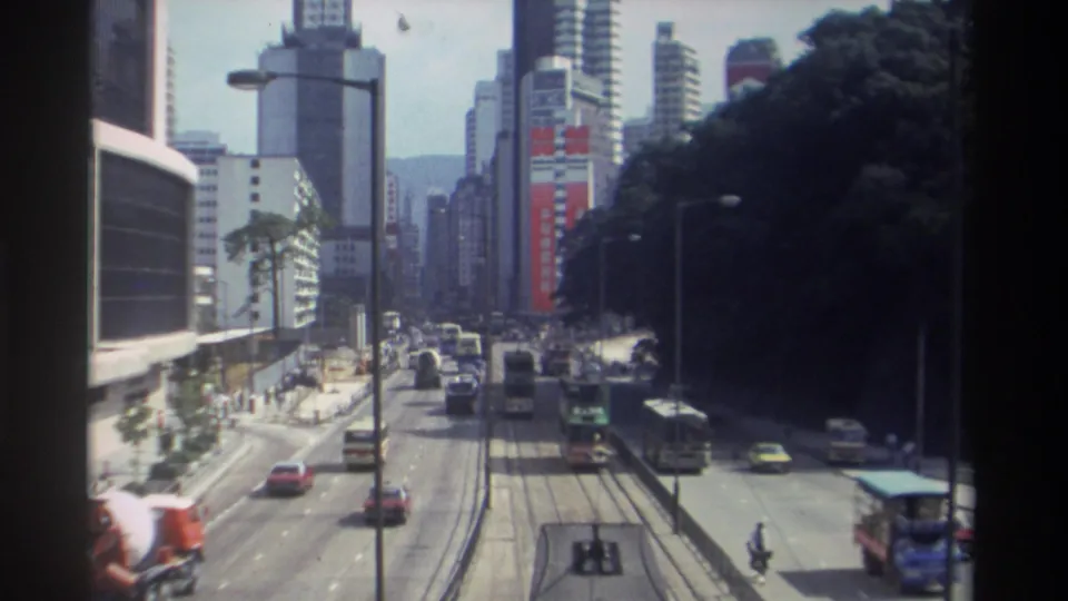 1982 Hong Kong elevated street view with buses, cars, tram rails, tall commercial buildings, and a distant person near the roadway.