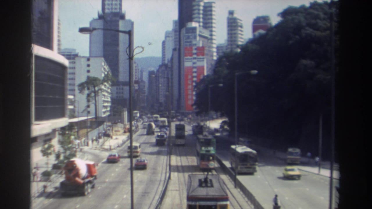 1982 Hong Kong home-movie frame showing multi-lane traffic below high-rises, with double-decker transit vehicles and distant hills.