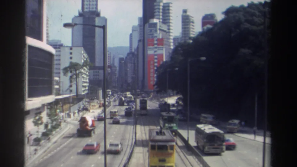 Elevated 1982 Hong Kong street view with mixed traffic, double-decker transit, tall buildings, signage, and a wooded slope at right.