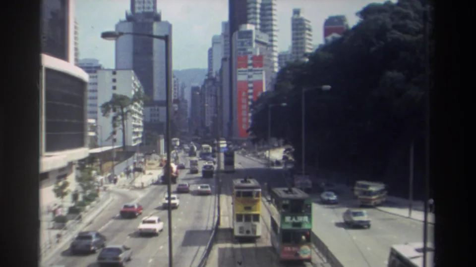 Late clip frame showing 1982 Hong Kong traffic from above, including double-decker transit, cars, tall buildings, signage, and hillside greenery.