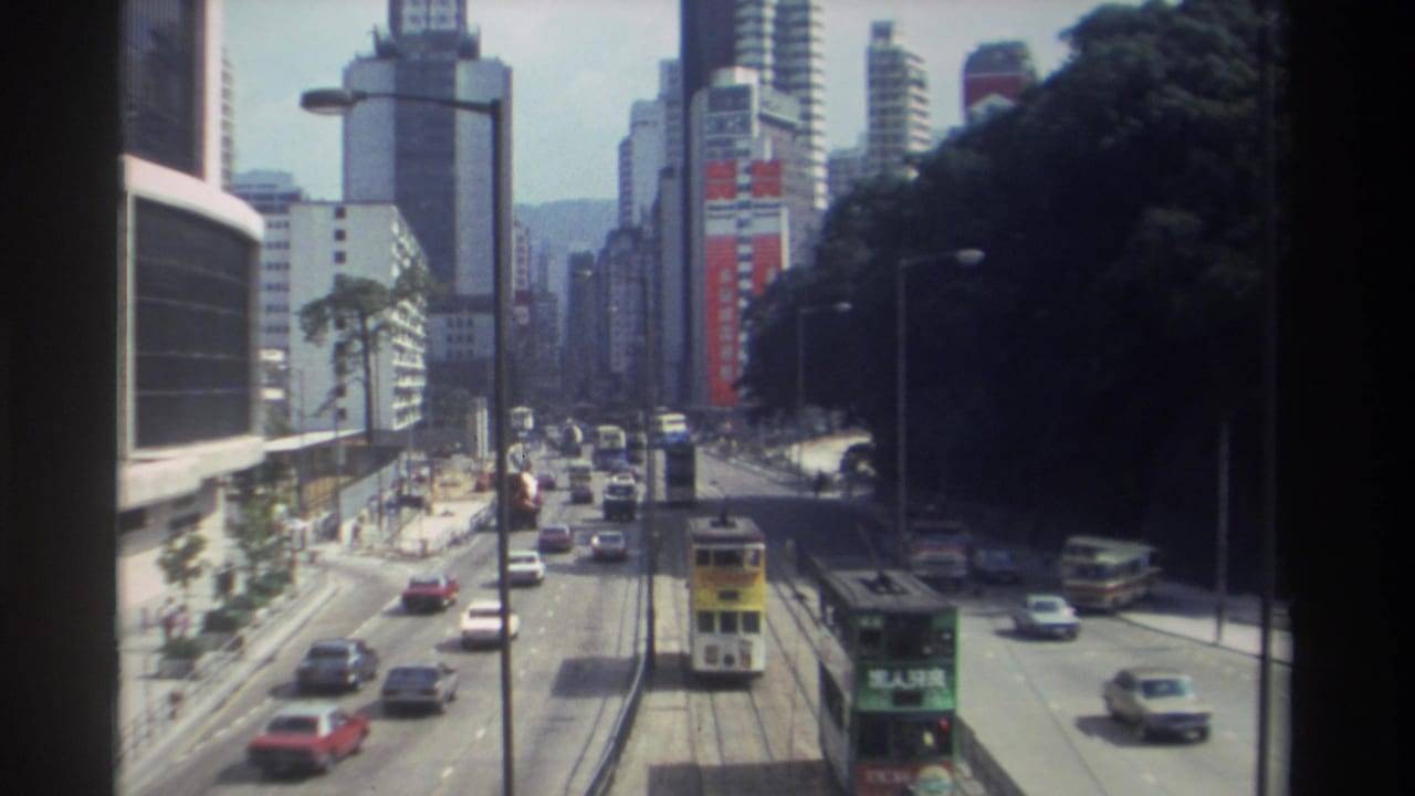 1982 Hong Kong street traffic seen from above, with tram tracks, cars, buses, high-rises, trees, and advertising panels.