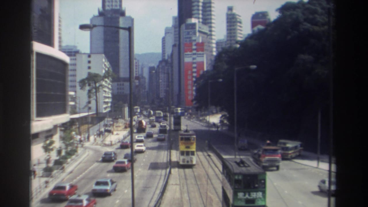 Elevated 1982 Hong Kong view showing a yellow tram, a green tram, cars, buses, roadside trees, and high-rise facades.