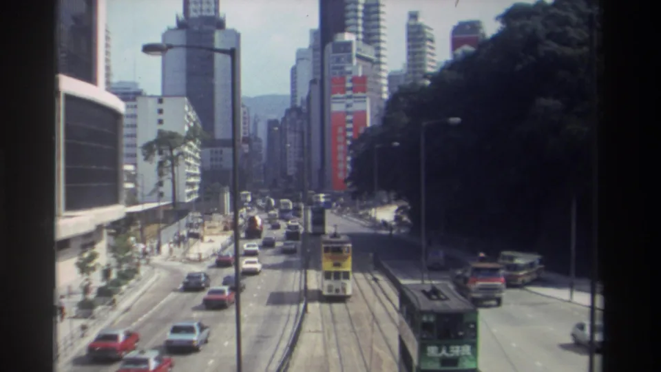 Elevated 1982 Hong Kong view showing a yellow tram, a green tram, cars, buses, roadside trees, and high-rise facades.