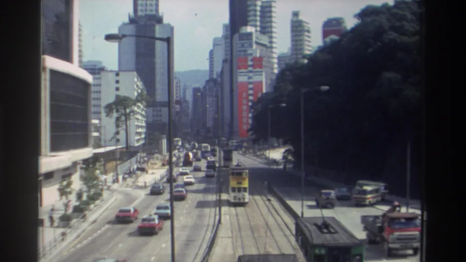 Hong Kong traffic in 1982, viewed from above with tram rails, a double-decker tram, cars, buses, pedestrians, and dense buildings.