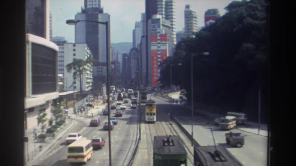 1982 Hong Kong elevated street view with a double-decker tram, bus, cars, high-rise buildings, trees, and visible advertising.