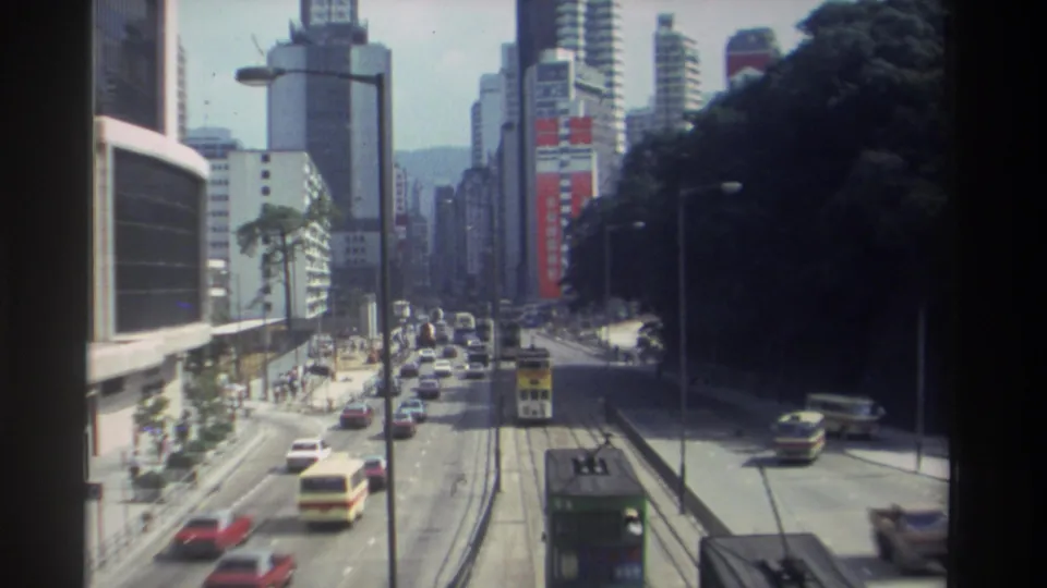 1982 Hong Kong elevated street view showing traffic lanes, tram tracks, double-decker vehicles, hillside greenery, and dense downtown high-rises.