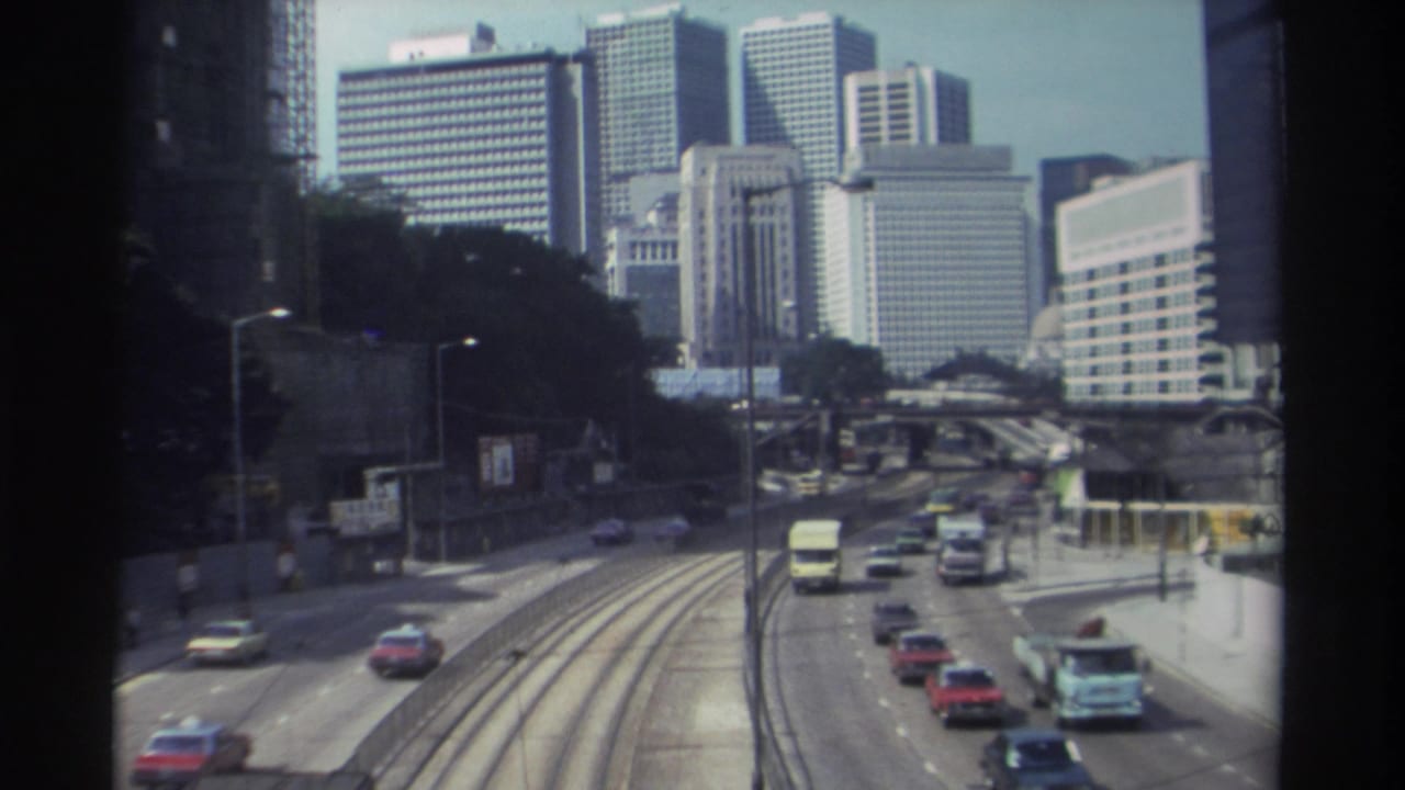 1982 Hong Kong roadway scene with curving tram rails, moving traffic, roadside greenery, and a cluster of downtown high-rise buildings.