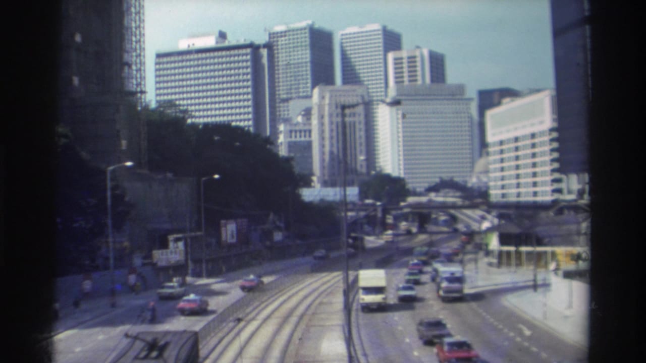 1982 Hong Kong street traffic passes tram-lined lanes beside a wooded slope and tall commercial buildings viewed from above.