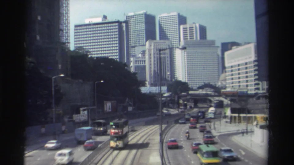1982 Hong Kong home-movie frame of mixed road traffic, tram tracks, and tall commercial buildings seen from above.