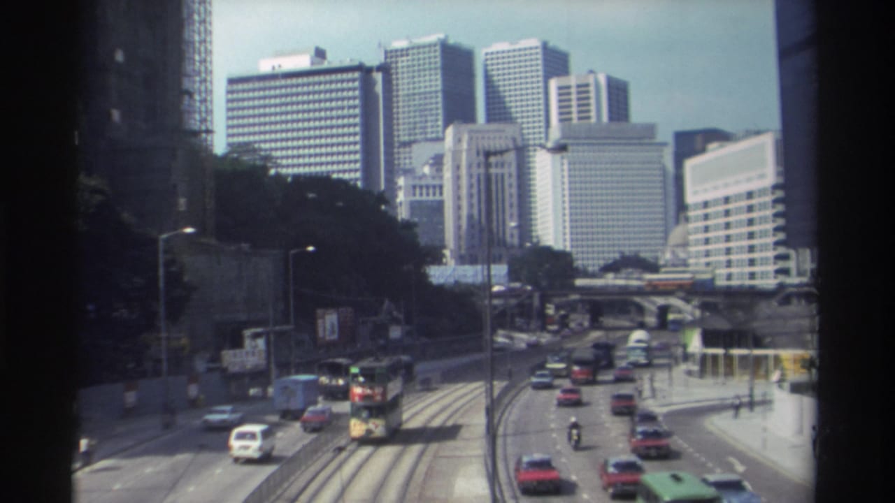 1982 Hong Kong home-movie view of a double-decker bus and cars moving along tram tracks below a cluster of high-rise buildings.