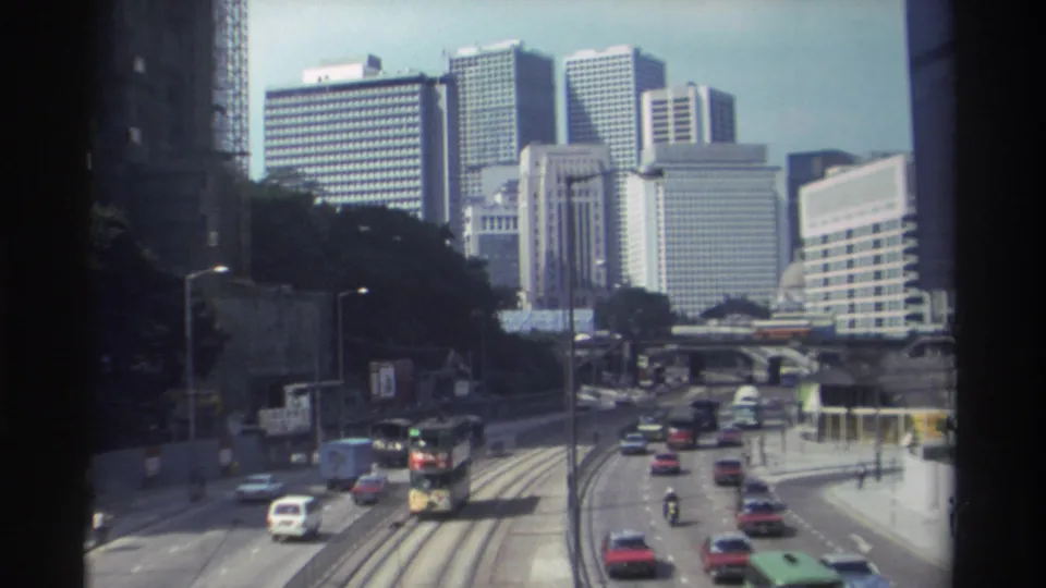 1982 Hong Kong home-movie view of a double-decker bus and cars moving along tram tracks below a cluster of high-rise buildings.