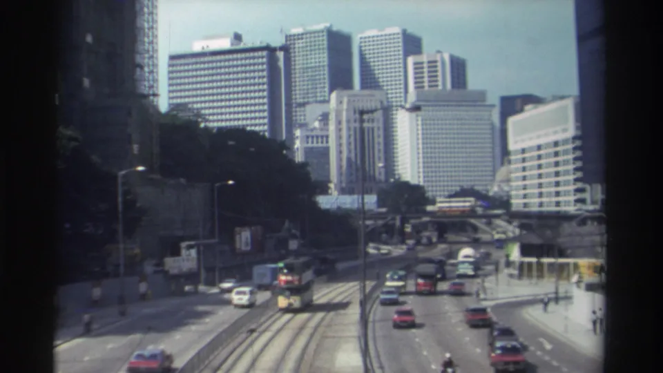 Elevated 1982 Hong Kong street view with vehicles moving around tram tracks and a double-decker bus under a dense high-rise skyline.