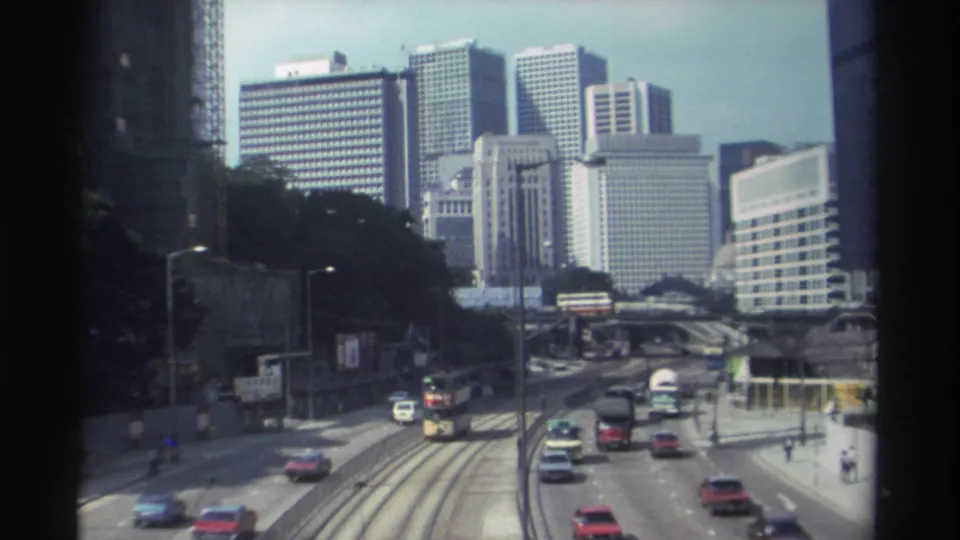1982 Hong Kong elevated traffic shot showing a bus near tram tracks, surrounding cars, roadside trees, and tall commercial buildings.