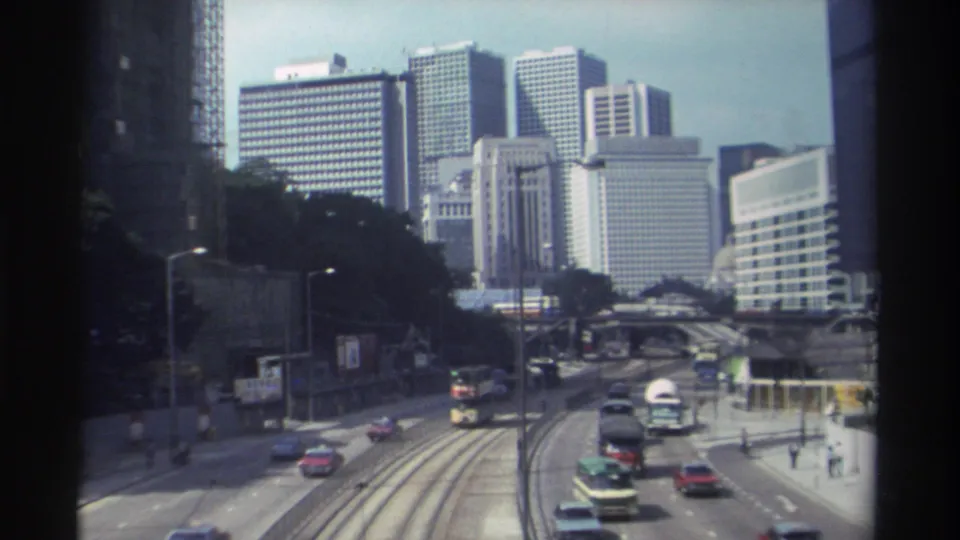 Hong Kong 1982 home-movie frame of multilane traffic with a double-decker bus, tram tracks, pedestrians, trees, and high-rise buildings.