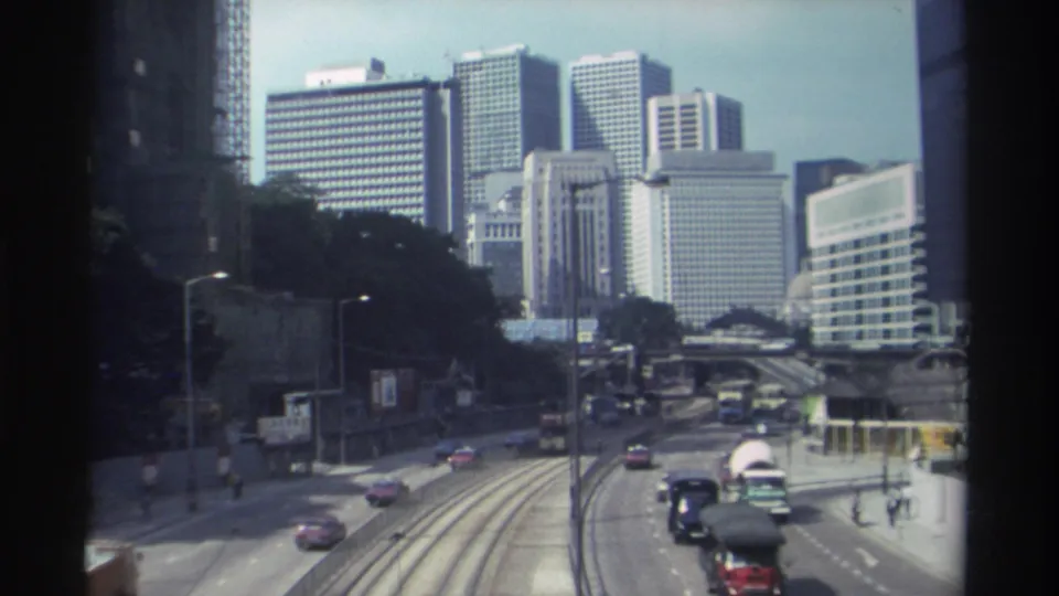 Elevated 1982 Hong Kong view with cars, buses, tram tracks, roadside pedestrians, and clustered high-rises under a hazy sky.