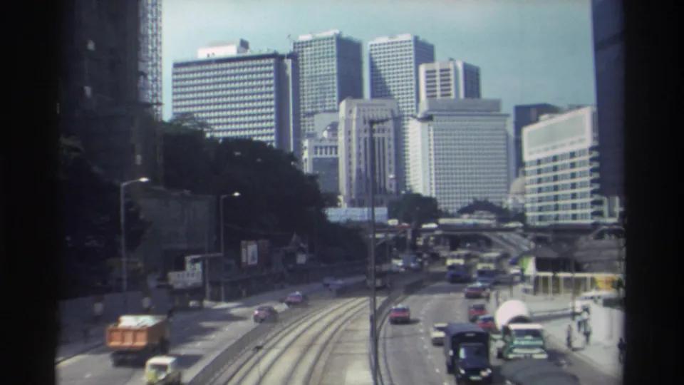 1982 Hong Kong traffic scene with a bus, cars, visible tram rails, small pedestrians, and modern high-rise blocks in the distance.