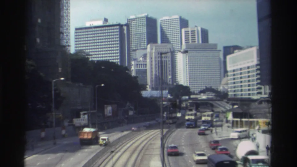 Late clip frame of 1982 Hong Kong traffic, showing cars, buses, tram tracks, pedestrians, and a dense high-rise skyline.