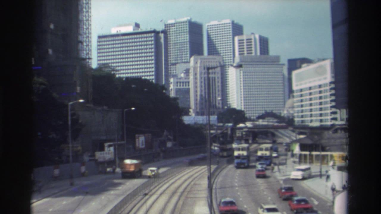 1982 Hong Kong roadway viewed through a window-like frame, with cars, buses, elevated roads, and high-rise buildings in the distance.