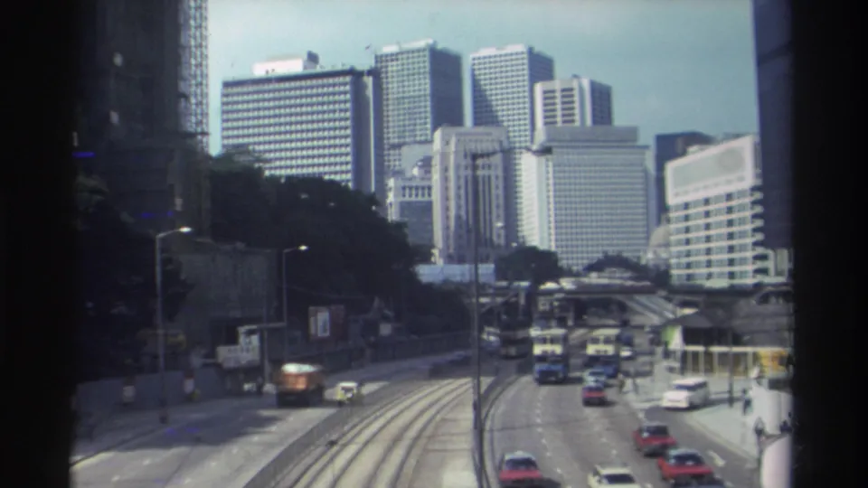 1982 Hong Kong roadway viewed through a window-like frame, with cars, buses, elevated roads, and high-rise buildings in the distance.