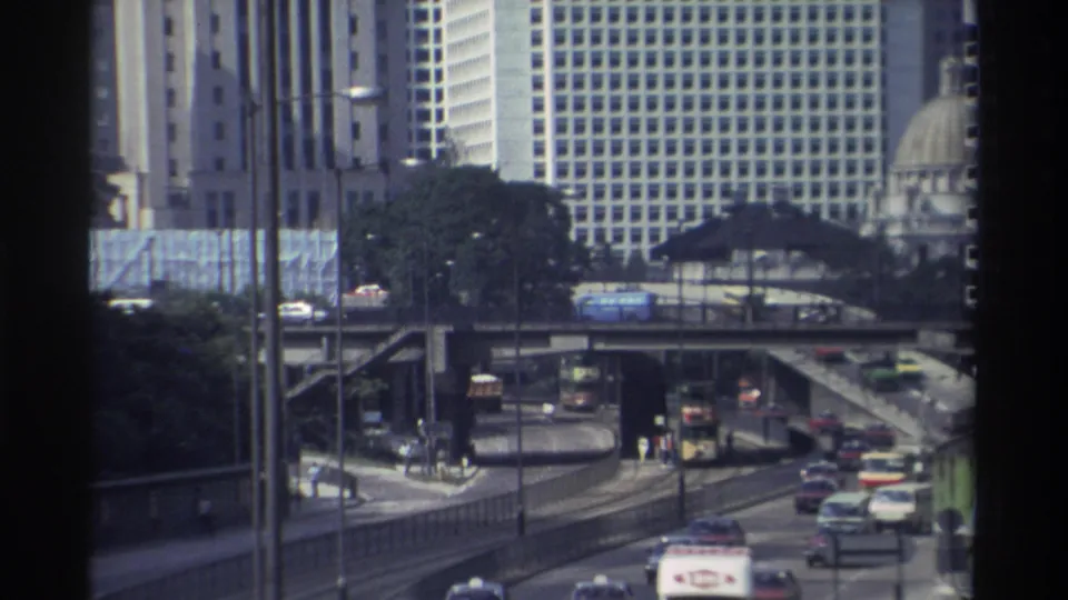 1982 Hong Kong traffic scene with elevated overpasses, cars, double-decker buses, high-rise buildings, and a distant domed structure.
