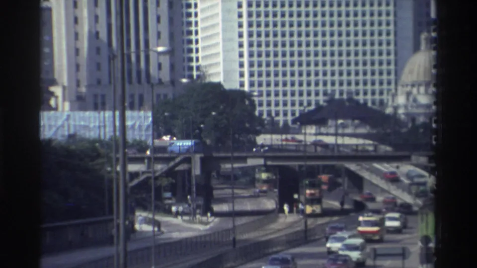 1982 Hong Kong street view showing buses, cars, pedestrians near crossings, elevated roads, trees, and high-rise buildings.