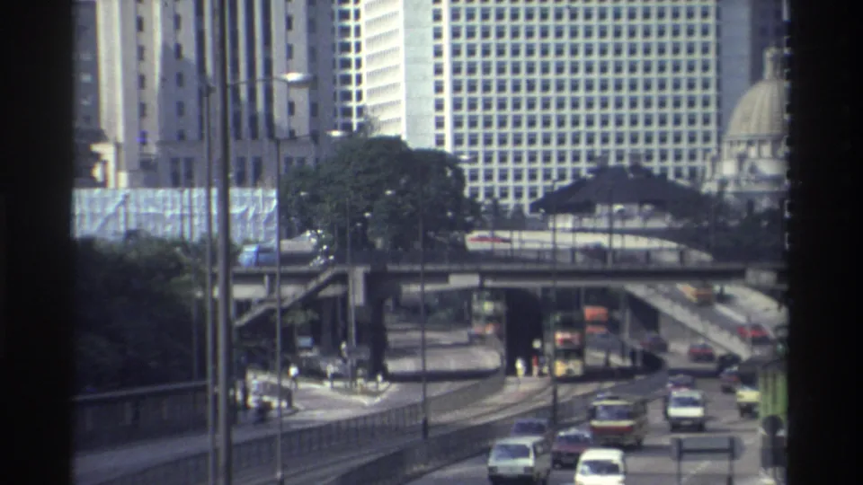 1982 Hong Kong traffic moving along a divided road under an overpass, with buses, cars, pedestrians, trees, and high-rises visible.