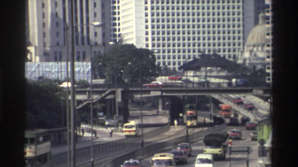 1982 Hong Kong roadway view with a bus, small cars, and a motorbike moving beneath a pedestrian bridge and high-rises.