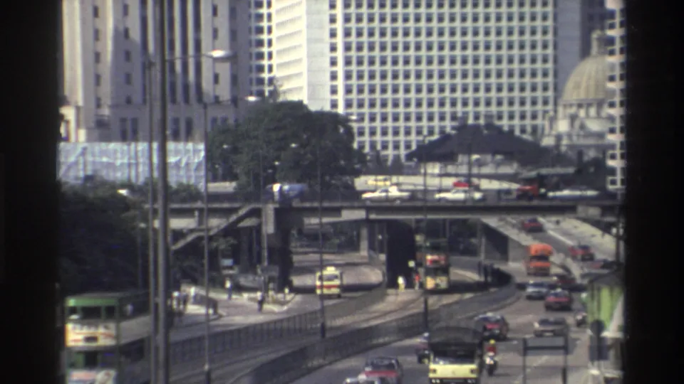 Hong Kong traffic in 1982 continues through multi-level roads, with buses, cars, and high-rise buildings in soft 8mm footage.