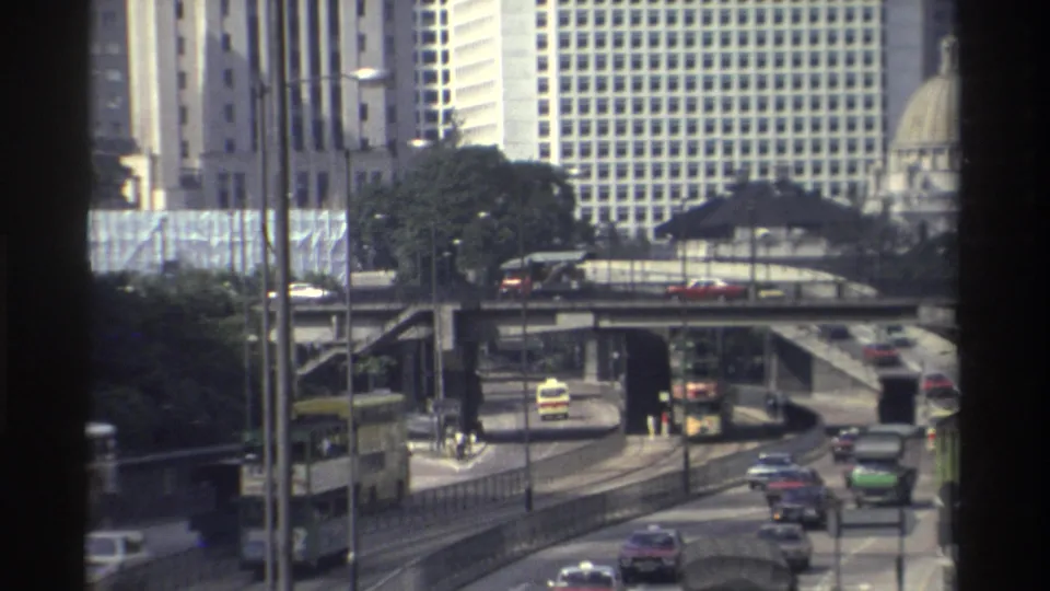Hong Kong traffic in 1982 passes through elevated downtown roads, with buses, cars, trees, and tall office buildings visible beyond.