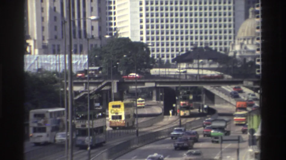 1982 Hong Kong street traffic with double-decker buses, sedans, trucks, elevated lanes, and high-rise buildings filmed from behind a window edge.