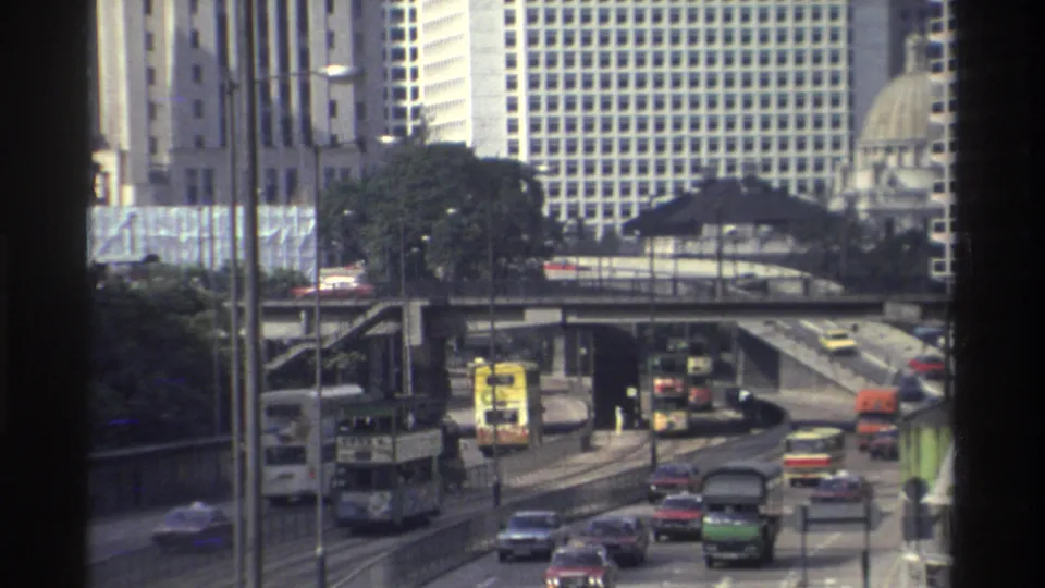 End frame of 1982 Hong Kong downtown traffic, showing buses, cars, trucks, elevated roads, and high-rise buildings through a window-framed view.