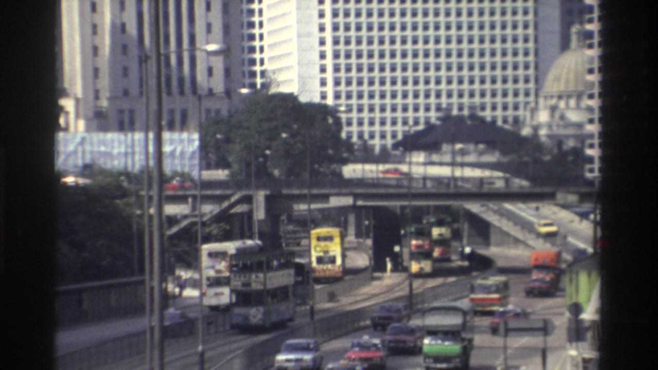 Hong Kong street traffic with double-decker buses, cars, elevated roads, and high-rise buildings.