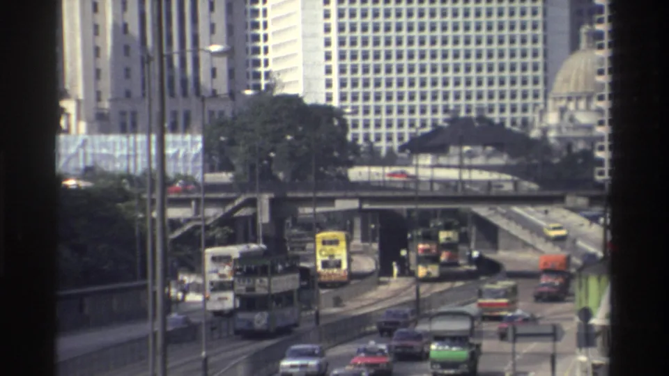 1982 Hong Kong traffic scene viewed through a window, with double-decker buses, cars, overpasses, towers, and a domed roofline.