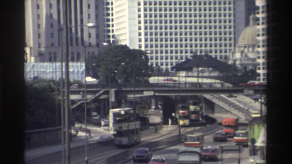 Hong Kong urban roadway in 1982 with double-decker buses, cars, elevated ramps, trees, and gridded high-rise facades.