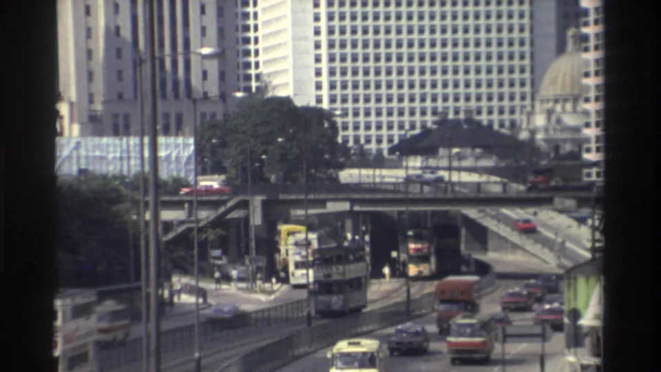 Hong Kong traffic in 1982 with a double-decker bus, cars, an orange truck, elevated roadway, and tall city buildings.