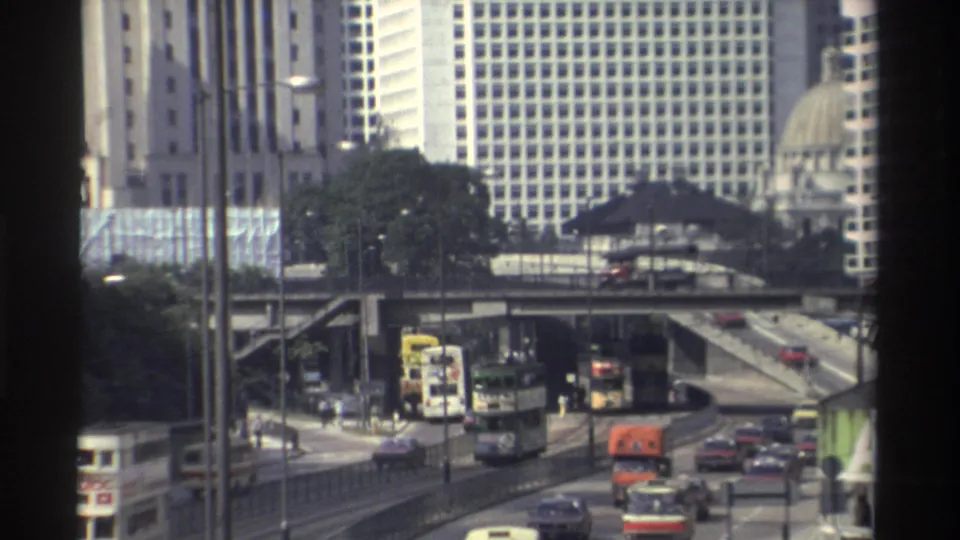 1982 Hong Kong traffic seen from behind a dark frame, with buses and cars moving beneath high-rise buildings and a distant dome.