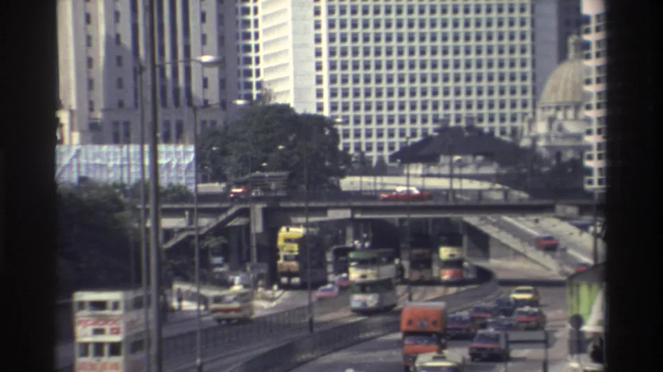 1982 Hong Kong street traffic with buses, taxis and trucks on layered roads, framed by high-rise buildings and a dark vertical edge.