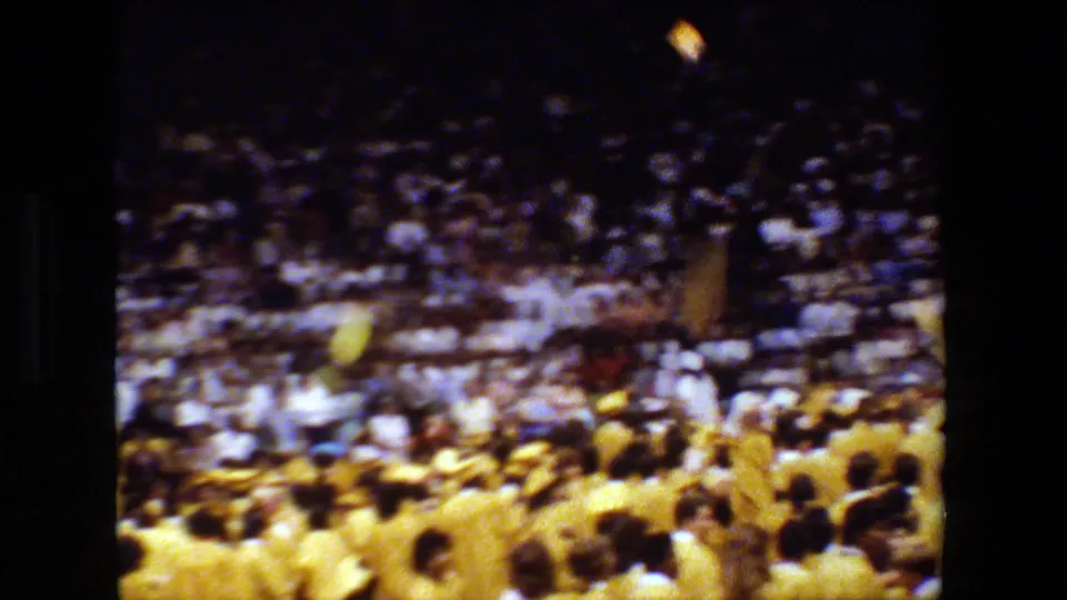 1983 Lansing-area graduation scene with graduates in yellow caps and gowns raising and throwing caps in a packed stadium.