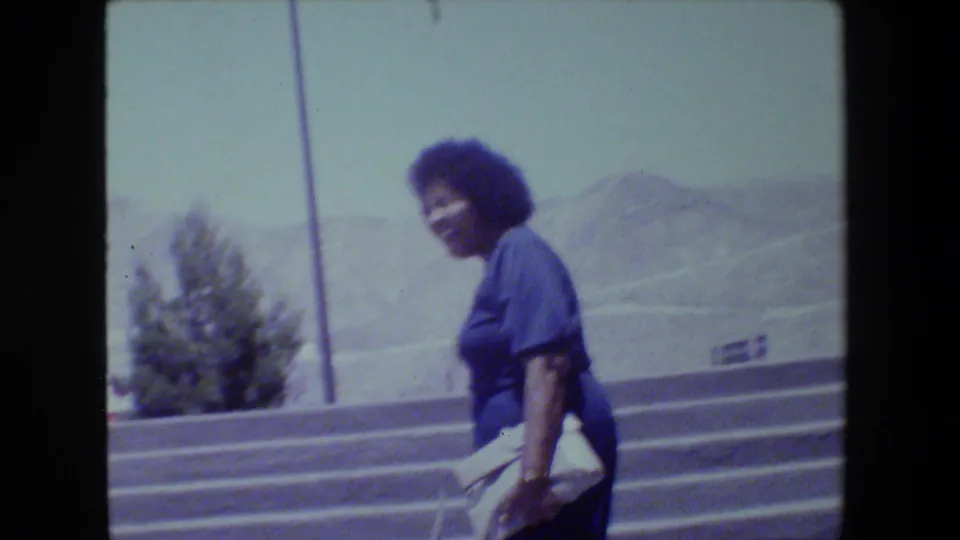 Home-movie frame of a woman in a blue dress walking down steps and holding a light-colored clutch or book, with a hazy landscape behind.