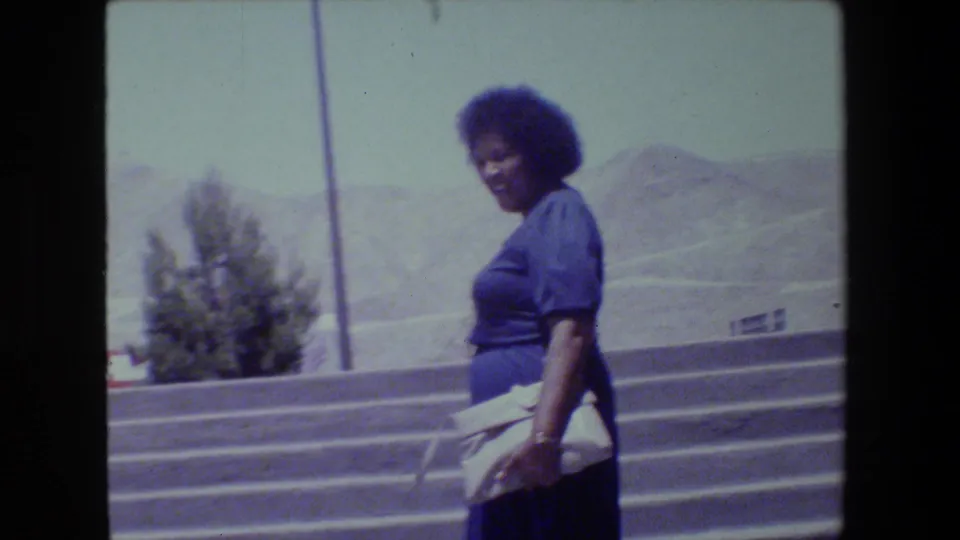 Woman in a blue dress pauses or turns on outdoor steps, smiling toward the camera against trees, pavement, and distant hills.