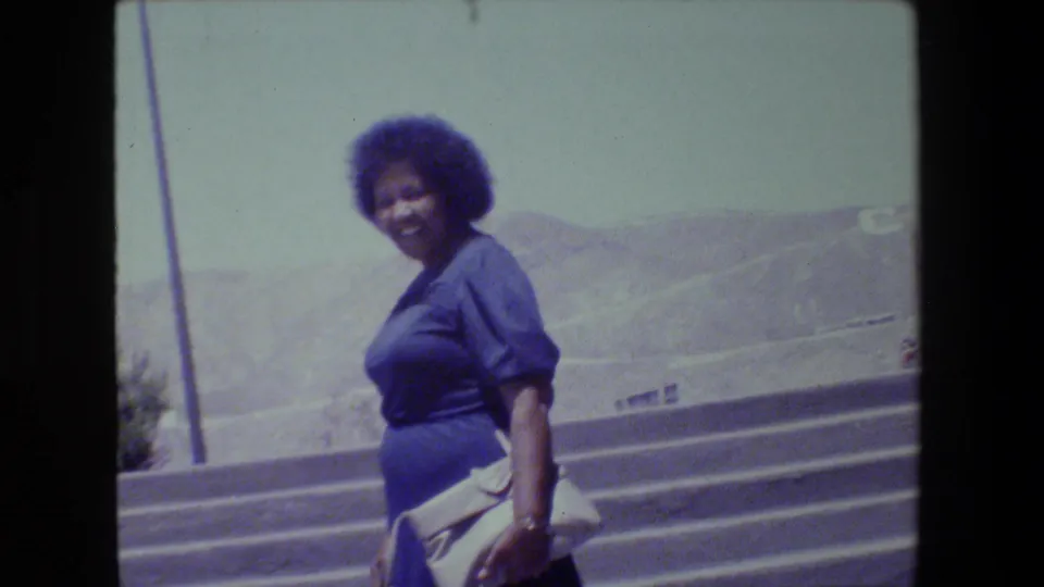 In supported 1983 Lansing home-movie footage, a woman in a blue dress smiles toward the camera while climbing outdoor concrete steps.