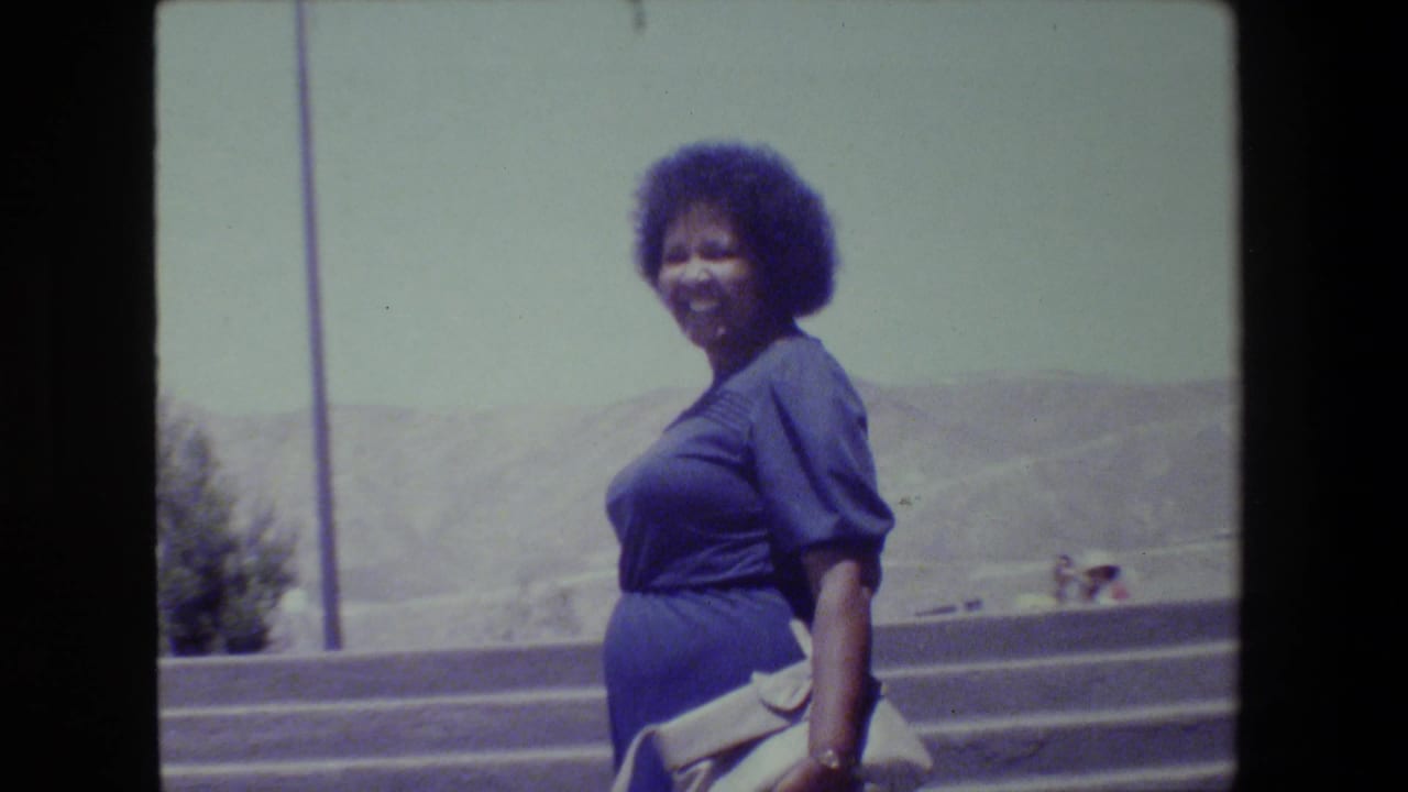 A woman in a blue dress walks up outdoor concrete stairs in the 1983 Lansing clip, with a guardrail and hazy hills behind her.