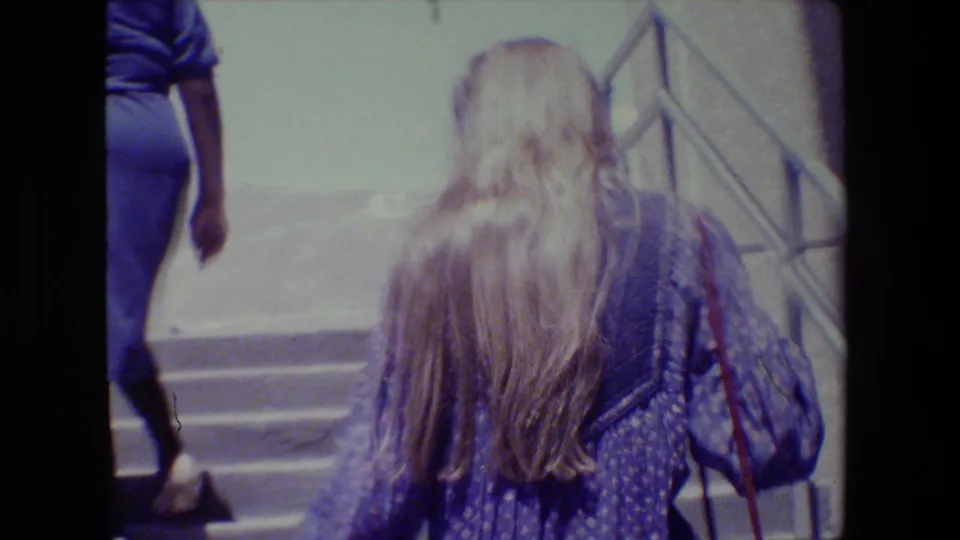 A younger long-haired blonde woman in a patterned top follows a woman in a blue dress up outdoor concrete stairs in the home-movie frame.