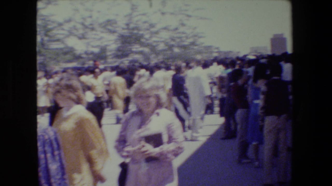 Crowd of adults walking outdoors on a sunny walkway with trees and buildings in the distance.