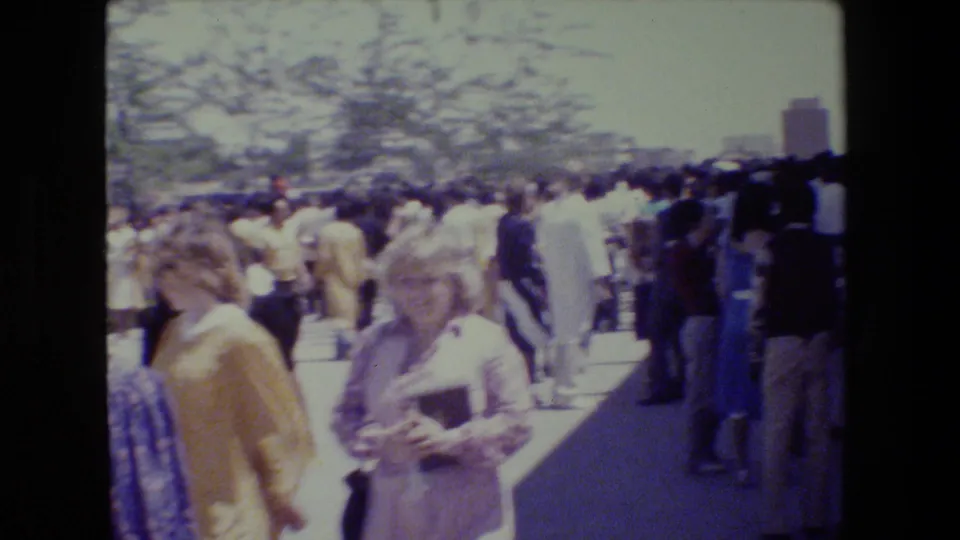 1983 Lansing home-movie frame showing adults gathered outdoors near a sunlit walkway, with trees and distant buildings behind them.