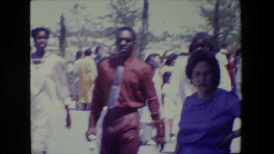 1983 Lansing church-event footage shows adults walking past the camera in dress clothing during a crowded outdoor gathering.
