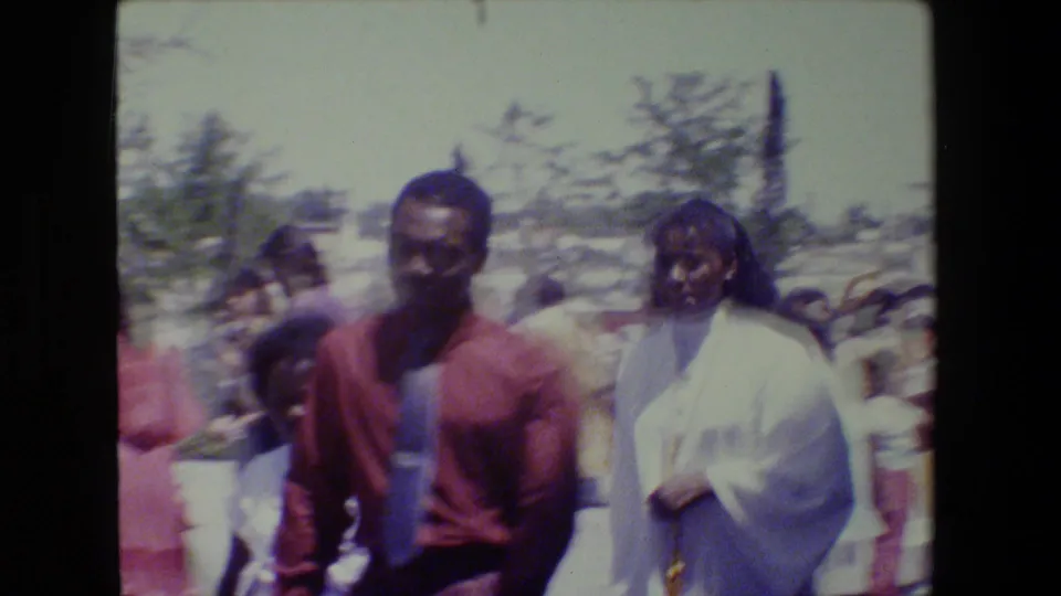 A handheld 8mm frame from Lansing in 1983 shows adults in dress clothing walking through a sunny outdoor church gathering.