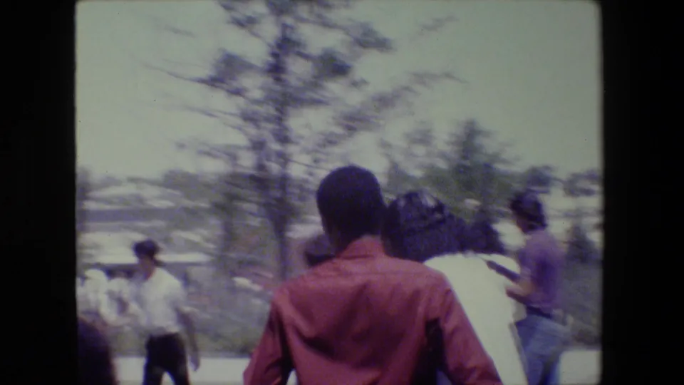 The closing 1983 Lansing frame shows people from behind as they move through the outdoor gathering, with trees and a bright sky in view.